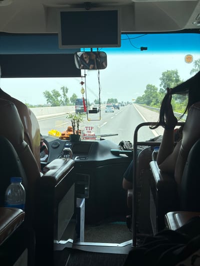 The image captures the interior of a bus or coach, looking forward through the windshield onto a road. The perspective is from a passenger seat, with a person's arm and long dark hair visible in the foreground, seemingly holding a phone. A plastic water bottle rests next to the seat.
The dashboard features various controls, a built-in screen, and a rearview mirror reflecting a person's face. Decorative items are placed on the dashboard, including a small potted plant, a golden Buddha statue, and red and gold hanging ornaments. A white sign with prominent red text reads "TIP FOR BUS DRIVER & TOUR GUIDE."
To the left, a monitor is mounted, and a dashcam or small camera is attached to the upper part of the windshield. A circular sticker or permit is also affixed to the windshield, showing numbers that are partially legible (e.g., "105030").
Beyond the windshield, a multi-lane highway or main road stretches into the distance, with several cars and trucks visible, indicating traffic. The weather appears clear and sunny, with a bright blue sky and green trees lining the road. The scene suggests daytime travel on a busy road in Hải Phòng, Vietnam.