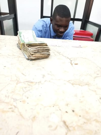 A man is behind a counter, apparently counting a large stack of Nigerian 500 Naira notes. The scene appears to be inside a bank, possibly in Satellite Town, Nigeria. He wears a light blue dress shirt and a patterned tie. Red file folders are stacked behind him to his right. A rubber band secures the stack of bills. Bank forms are on the counter to the right of the money. The counter is marble. A dark frame surrounds the counter area.
