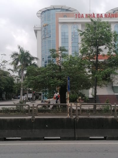 The image shows a street scene in Ha Tu Ward, Vietnam. A large building with a curved glass facade and the words "TOA NHA DA NANG" on its roof dominates the background. A palm tree and other trees are interspersed between the building and the viewer. A metal fence and concrete barriers are situated along the roadside in the foreground. A person carrying balloons walks behind the fence and in front of a tree. The sky is overcast.
