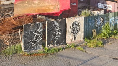 In Södertälje, Sweden, three concrete slabs stand vertically, each adorned with elaborate black and white graffiti art resembling intricate, stylized skulls or alien-like heads. The slab on the left features a detailed, dark, swirling pattern with prominent eye sockets. The middle slab shows a similar, flowing design with a prominent, multi-faceted eye. The rightmost slab presents a more distinct, mechanical-looking skull with a central eye-like aperture. These artworks are positioned on the edge of a paved area, with a grassy verge and weeds growing around their bases. Behind the concrete slabs, a corrugated metal fence, also marked with graffiti, runs horizontally. Further back, a large red industrial container or truck bed is visible, along with piles of rusted metal mesh and other industrial debris. The scene appears to be an outdoor industrial or junkyard area.