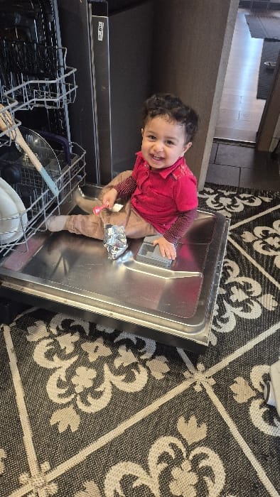 An indoor scene features an infant seated on the fully extended lower rack of an open dishwasher. The infant, possessing dark curly hair and light brown skin, is dressed in a red collared shirt with dark long sleeves underneath and light brown pants. The infant is smiling and looking directly at the camera. In the infant's right hand, a small pink utensil is held. On the stainless steel dishwasher rack, adjacent to the infant's right leg, a crumpled piece of aluminum foil is visible. To the infant's left, a gray plastic component of undetermined function rests on the rack. The dishwasher's upper rack contains various dishes and utensils, including a wooden spoon, a pot lid, and plates. The floor is covered by a rug with a repeating black and off-white floral/damask pattern. In the background, a dark cabinet or wall is present, alongside a doorway leading to an adjacent room with a lighter-toned floor. The specific location within the city of Melle, Belgium, cannot be determined from the provided image.