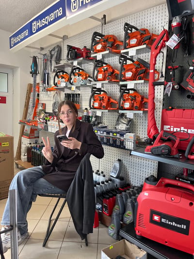 Here's a detailed description of the image:

**Location and Environment:** The image appears to be taken inside a store that sells power tools, possibly in the city of Rudozem, Bulgaria. This is suggested by textual information extracted from the image with OCR. The background shows walls covered with various tools on display, including chainsaws, leaf blowers, and hedge trimmers.

**People:** A young woman is seated on a black metal chair in the middle of the frame. She is wearing glasses, jeans, a brown jacket, and white sneakers. She is holding a cell phone in one hand and making a peace sign with the other.

**Objects:** Besides the various tools mentioned, there are oil bottles, boxes with labels, and a sign displaying "Husqvarna." The woman is holding a black cell phone.

**Action and Interactions:** The woman is posing for a picture, as suggested by her hand gesture and phone positioning.

**Forensic details:** The chainsaws are prominently displayed and are likely a main product of the store. The brands visible are Husqvarna and Einhell. The products appear well-organized and ready for sale.