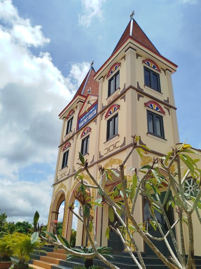 An exterior, low-angle, medium shot captures a tall, cream-colored building with a red, tiled, pointed roof, set against a bright blue sky with scattered white clouds. The building features multiple levels with dark-framed windows and decorative colorful arches above some of them. A sign on the building reads "ĐẠI ĐẠO TAM KỲ PHỔ ĐỘ THÀNH THẤT XUÂN SƠN" in blue lettering on a white background. The lower portion of the building has arched doorways and a set of stairs leading up to them. In the foreground, leafless trees with green leaves and a few pink flowers are obscuring parts of the building. Lush green foliage and palm trees are visible to the left and behind the building, suggesting an outdoor setting in Xuân Sơn, Vietnam. There are no people visible in the image.