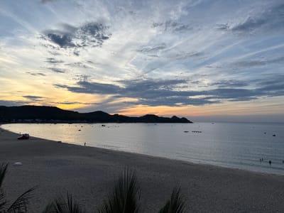 This is a panoramic, eye-level, outdoor photograph of a beach at sunset in Lệ Uyên, Vietnam. The sky is filled with dramatic clouds in shades of grey, white, and light blue, with streaks of yellow and orange near the horizon, indicating the setting sun. The sea is calm, reflecting the colors of the sky. The beach is wide and sandy, with a few palm fronds in the foreground obscuring the very bottom of the frame. In the distance, a dark, hilly coastline is visible, dotted with buildings. Several small boats are scattered across the water. On the beach, a red vehicle with a trailer is parked near the water's edge, and a few people are visible, including one walking on the shore and several others swimming in the sea. The overall scene is serene and tranquil.