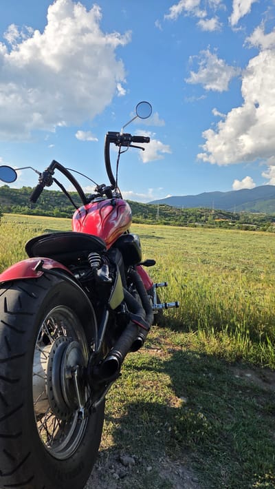 A red and yellow flame-painted motorcycle is parked on a dirt path bordering a grassy field. The motorcycle is viewed from the rear, slightly to the left, with its prominent exhaust pipes and rear tire in focus. Beyond the field, rolling hills dotted with trees and some power lines form the background, under a bright blue sky with scattered white clouds. The scene is set in the Rila region of Bulgaria, evidenced by the natural landscape. There are no people visible in the image.