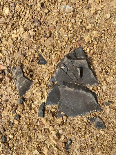 A close-up, top-down view shows dark, fragmented rock pieces scattered on a sandy, gravelly ground. The rock fragments are angular and appear to have sharp edges, with one large piece in the center exhibiting visible cracks and a slightly raised protrusion. The surrounding ground is composed of a mixture of small, irregularly shaped pebbles and sand, varying in color from light tan to reddish-brown and grey. The lighting suggests an outdoor environment, casting subtle shadows on the ground. There are no people or discernible structures visible in the image. The location is the city of Ko Siboya, Thailand.