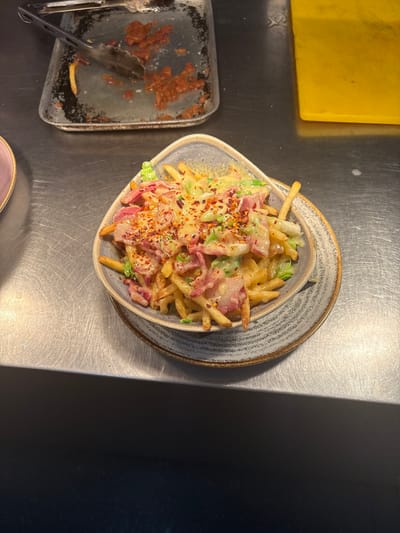 The image shows a plate of french fries covered in a white sauce, bacon bits, green onions, and red pepper flakes. The plate is on a stainless steel table in a restaurant. The plate has a gray and blue pattern, with a textured design. The image is taken from a slightly elevated angle. The restaurant appears to be in Newton upon Derwent, United Kingdom. The coordinates are: 53.936, -0.904. There are no people in the image.