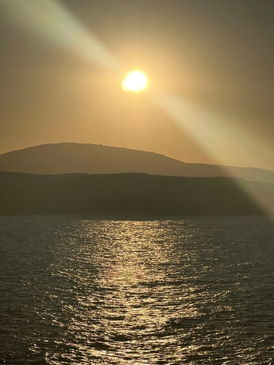 The image captures a serene sunset over the sea near Sinemorets, Bulgaria. The sun, a bright orb, is partially obscured by hazy atmospheric conditions, casting a golden light across the sky and water. Prominent, silhouetted mountain ranges form the background, with layers of hills receding into the distance. The sea in the foreground is rippled, reflecting the warm hues of the sunset, creating a shimmering effect. The overall scene evokes a sense of tranquility and natural beauty, typical of coastal landscapes at dusk.