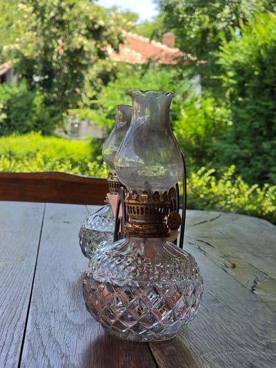 Two antique oil lamps are placed on a wooden table outdoors. The lamps have clear, cut-glass bases and metal accents. In the background, lush green trees and a building with a tiled roof are visible, suggesting a rural or suburban setting. The lighting indicates it is daytime, likely with sunlight filtering through the trees. The image appears to be a close-up shot, focusing on the lamps and the texture of the wooden table. The location is Indzhe Voyvoda, Bulgaria.