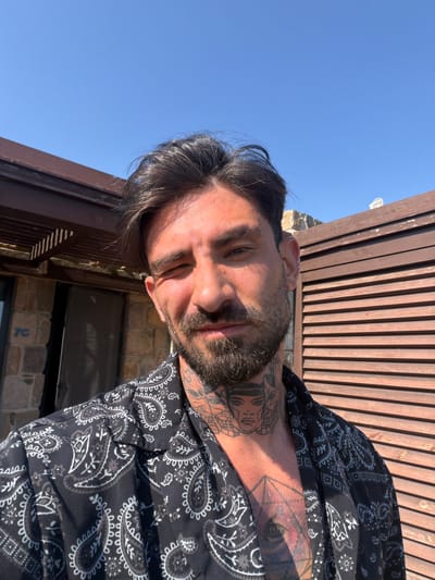 A man is taking a selfie outdoors in front of a wooden structure and a stone building under a clear blue sky. The man has dark hair, a beard, and tattoos on his neck and chest. He is wearing a black shirt with a white paisley pattern. The building behind him appears to be a resort or hotel, with a numbered "7C" visible on the stone wall. The overall setting suggests a sunny, warm climate, likely in a tourist area.