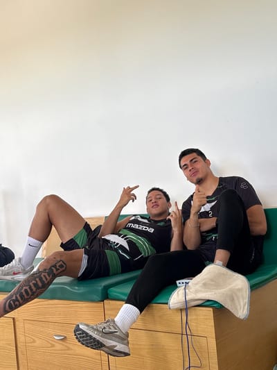 Two males are depicted seated on a green-cushioned wooden bench within an indoor room in Las Crucitas, Mexico. The background consists of a plain white wall.
The individual on the left, with dark hair and light skin, is reclined on the bench. He is wearing a black sleeveless athletic top with green and white design elements, including a 'Mazda' logo. He wears black shorts with green and white stripes, white Nike socks, and white athletic shoes with red accents. A large, dark-inked tattoo is visible on his right thigh. His left arm is bent, and his hand is raised, displaying an extended index finger and pinky finger gesture. His right hand rests near his abdomen.
The individual on the right, also with dark hair and light skin, is seated more upright on the bench. He is wearing a black short-sleeved athletic shirt, black athletic pants, and white socks. A thin gold-colored bracelet is visible on his left wrist. His right foot is enclosed within a tan-colored padded boot or wrap from which blue wires emerge. His left arm is bent, and his hand is raised, displaying an extended index finger and pinky finger gesture. His right arm rests on his lap.
Both males are looking towards the camera. The wooden structure supporting the green bench cushion contains multiple drawers equipped with silver-colored pull handles.