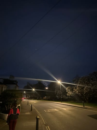 The image depicts a nighttime street scene in Salisbury, United Kingdom. A lone individual is walking away from the viewer on a paved sidewalk along the left side of the frame. The person is an adult, observed from behind, wearing a dark top with red upper panels and dark trousers, carrying a dark bag on their left shoulder. A row of dark bollards with brass-colored tops separates the sidewalk from the asphalt road.
The road curves gently to the right and is illuminated by several streetlights, one prominent in the mid-right foreground exhibiting significant lens flare. Further down the road, a vehicle with illuminated red taillights is visible, moving away from the camera. On the left, partially obscured by bushes and trees, a multi-story building with a light-colored facade is visible.
To the right of the road, a grassy verge leads to a line of trees, one with light-colored, possibly flowering foliage, stands out under the streetlight. In the sky above the tree line and behind other trees, a faint, circular moon is visible through light cloud cover. The dark blue night sky occupies the upper portion of the frame, crisscrossed by four distinct parallel power lines running diagonally from upper left to lower right. A single bright celestial body, possibly a star or planet, is visible in the upper-mid section of the sky. The overall scene is lit primarily by artificial street lighting.
