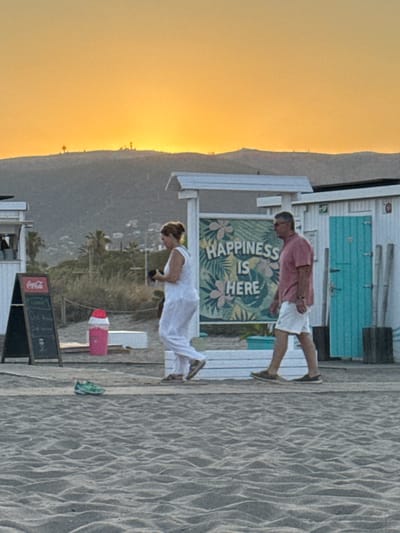 The image depicts a beach scene at sunset in Castelldefels, Spain. Two individuals are walking along the sandy shore. A woman in a white outfit is holding a dark object in her hands, while a man in a pink shirt and white shorts walks beside her. Behind them, a sign with the text "HAPPINESS IS HERE" is visible, surrounded by tropical-themed graphics. To the left, a Coca-Cola advertisement is displayed on a chalkboard easel. Further in the background, a pink trash can with a red lid is situated near some white structures. The sunset casts a warm, orange glow across the sky, illuminating the hills in the distance. A single green shoe lies on the sand in the foreground. The overall environment suggests a relaxed, coastal atmosphere typical of a Spanish beach town.
