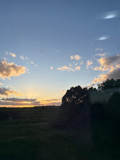 The image captures a sunset scene in Мажэйкаўскі сельскі Савет, Belarus. The sky is a gradient of blue, transitioning to orange and yellow as it nears the horizon, with scattered cumulus clouds illuminated by the setting sun. Sunlight streams through a gap in the clouds, creating rays that extend downwards. Along the horizon, trees and vegetation silhouette against the bright light. The foreground consists of a dark, undefined area, potentially a field or wooded area. There is no presence of people.