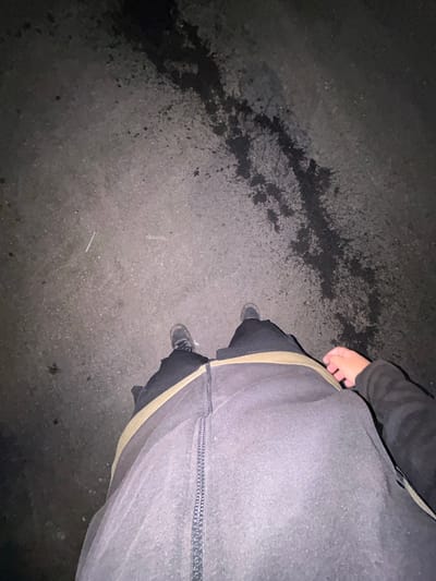 The image shows a first-person perspective of a person standing on an asphalt surface, likely a road or sidewalk, at night. The ground is dark with some lighter speckled areas and a dark, wet-looking stain in the upper portion of the frame. The person is wearing dark pants, a light-colored belt or waistband, and dark shoes. A hand with visible fingers is resting on the person's thigh. The lighting suggests an urban environment, and the detail about the location of Gorna Malina, Bulgaria, is not visually inferable from the image itself.