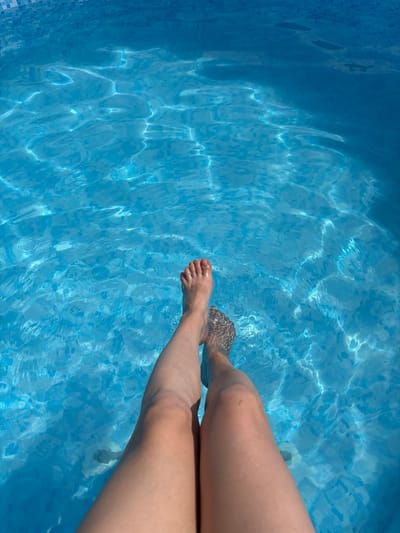 A high-angle medium shot shows a person's legs and feet submerged in a clear blue swimming pool. The water's surface is rippled, reflecting sunlight and creating bright, wavy patterns. The lower part of the frame is filled with water, and the upper part shows the edge of the pool, which is lined with small blue tiles. The person's feet are visible at the bottom of the frame, with one foot partially submerged and the other just touching the water. Their toenails are painted red. The overall impression is one of relaxation and refreshment. The specific location of Александровка, Russia, cannot be determined from the visual information provided in the image.