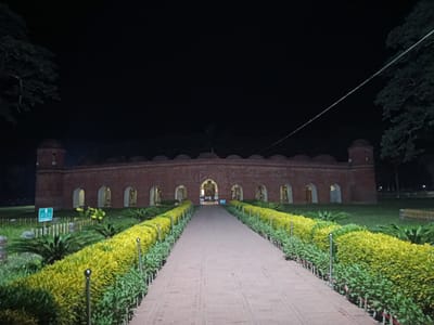 The image shows the Tomb of Khan Jahan Ali at night in Bagerhat, Bangladesh. A brick pathway, lined with manicured yellow and green bushes, leads towards the imposing brick structure. The building features a series of arched openings and two domed towers at either end. The sky is dark, and trees are visible on either side of the frame. The scene is illuminated by artificial lighting, casting a warm glow on the pathway and the building. There are no people visible in the image.