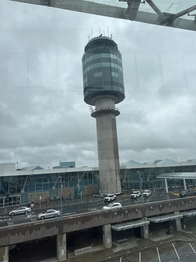 This media file captures an exterior view of a modern airport control tower and surrounding airport infrastructure under an overcast sky. The control tower, a prominent subject, features a cylindrical, glass-paneled observation deck with a checkered pattern of dark and light gray squares, rising from a sturdy concrete shaft. Adjacent to the tower, a multi-story airport terminal building with extensive glass facades is visible.

On the ground level, a tarmac area shows several parked vehicles, including white cars that appear to be airport security or service vehicles, and a darker colored vehicle. A section of what appears to be a parking garage or service area with numbered parking spaces (e.g., "23", "24") is also present. The ground surface is wet, suggesting recent rain.

The scene is viewed from a slightly elevated perspective, possibly from a window within another airport building or a nearby structure. The lighting suggests it is daytime, but the cloudy weather creates a diffused light, making it difficult to determine the exact time. There are no clear indicators of specific emotions, but the overall atmosphere is functional and typical of an active airport environment. No discernible text is clearly legible in the image. The location context is Richmond, Canada.