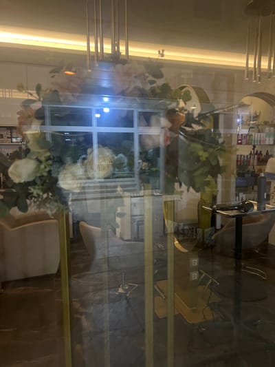 The image displays the interior of a hair salon or beauty parlor, viewed through a reflective glass partition in Smederevska Palanka Municipality, Serbia. The foreground is dominated by reflections on the glass, primarily featuring a decorative floral arch or frame composed of white and pink flowers with green foliage, supported by vertical gold-colored elements. Reflected in the upper center is a multi-paned window with bright blue illumination from beyond.

Within the salon, visible through the reflections, are various elements. On the right, shelves are stocked with numerous bottles of hair care products. A dark salon chair with a footrest and a small dark table holding a hairdryer and brush are present. A large, curved mirror with a gold frame is partially visible. On the left, light-colored upholstered chairs or seating are arranged on a dark floor. The ceiling features multiple hanging gold-colored cylindrical light fixtures and a continuous strip of warm yellow lighting along the perimeter. The environment appears modern and well-lit. No individuals are visible within the scene or its reflections.