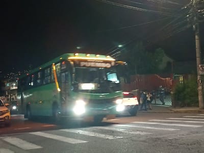 At night, a green bus with "PUEMBO" displayed is crossing a zebra crossing. Its headlights are on, illuminating the road. Behind the bus, a white car also has its lights on. Several people are standing on the corner near a red fence and gate. A street lamp and overhead power lines are visible. A "no parking" sign is posted on a pole on the right side. The background features houses on a hillside.