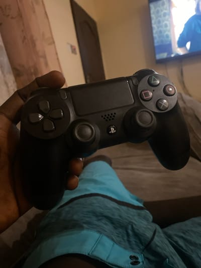 The image captures a close-up, angled view of a person's hand holding a black PlayStation DualShock 4 controller, set indoors in Lakka, Sierra Leone. The dark-skinned hand firmly grips the controller, with fingers visible wrapped around the bottom.

The controller is the primary focus, showcasing its distinct features. On the left side, the iconic face buttons—triangle, circle, cross (X), and square—are clearly visible, along with the directional pad below them. The two analog sticks are also prominent, with the left one partially obscured by the hand. The central touch pad, featuring the PlayStation logo (PS button) and a small speaker grille, is also distinct.

In the blurred background, a television screen displays bright, indistinct content, suggesting a video game or other media is being played or watched. To the right, part of a leg wearing blue and teal athletic shorts or sleepwear is visible, indicating the person is likely relaxing on a couch or bed. The overall lighting is dim, suggesting an evening or nighttime setting. The scene conveys a relaxed atmosphere of leisure and entertainment.