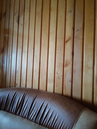 The image shows a close-up of a brown leather sofa in front of a wooden paneled wall. The wood paneling is light brown and oriented vertically, with visible grain and knots. The sofa has a prominent curved section with deep creases, creating a ribbed effect. The leather appears worn, with visible wrinkles and texture. The lower portion of the sofa, a lighter beige color, is also visible, with similar textured leather. The scene is indoors, with the lighting suggesting a daytime setting. There is no visible text in the image. The location context provided is Dubrovka, Russia, but there are no specific visual cues in the image to confirm this. The overall impression is of a simple, domestic interior.