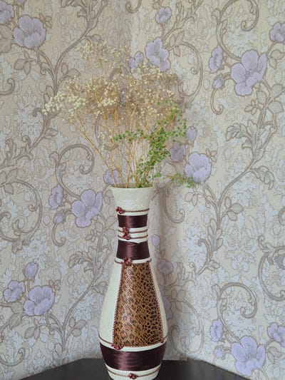 A decorative vase filled with dried flowers and greenery stands on a dark surface in front of a patterned wallpaper. The wallpaper features a repeating floral motif in muted tones of beige, white, and purple, with intricate scrolling vines and leaves. The vase itself is tall and shapely, with a cream-colored base and neck. It is adorned with dark brown satin ribbons and a central panel with a snakeskin-like texture. Small red floral embellishments are visible where the ribbons meet. The dried flowers inside the vase are delicate and pale, with some green foliage interspersed. The lighting is even, casting no harsh shadows. The scene appears to be an indoor still life, likely within a home environment.
