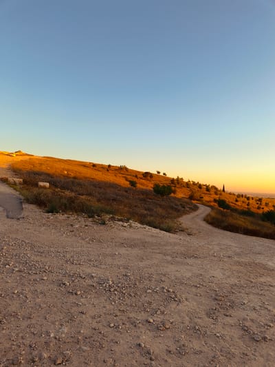 The image depicts a dirt path winding up a gently sloping hillside in Be'er-Sheva, Israel, during what appears to be sunset. The sky transitions from a clear, light blue at the top to a warm, golden orange near the horizon. The hillside is covered in dry, golden-brown vegetation, interspersed with sparse trees and shrubs. Several scattered, light-colored rocks are visible along the path and on the hillside. In the distance, a hazy view of a city can be seen. The path itself is composed of dirt and small stones, with some tire tracks visible in the foreground. The overall impression is one of a rural or semi-rural landscape bathed in the warm light of the setting sun.