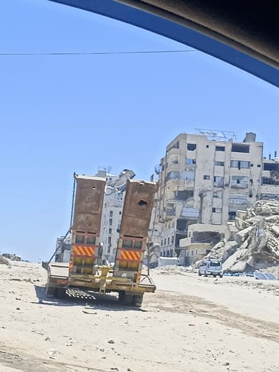 The image depicts a scene of destruction in Gaza. In the foreground, a damaged truck with a flatbed trailer is parked on a dirt road. The trailer is loaded with what appear to be metal beams or pipes. The truck's rear has red and yellow reflective markings.
In the background, several multi-story buildings are severely damaged, with visible structural damage, missing walls, and debris. One building in the center appears to be a residential or office complex, heavily impacted by shelling or other forms of destruction. Piles of rubble and debris are scattered around the base of the buildings, indicating the extent of the damage.
A white SUV is visible on the road in the mid-ground, driving away from the camera. The sky is clear and blue, suggesting it is daytime. The overall environment is dusty and arid, consistent with a desert or semi-arid climate. The scene conveys a sense of ongoing conflict and devastation in an urban area.
