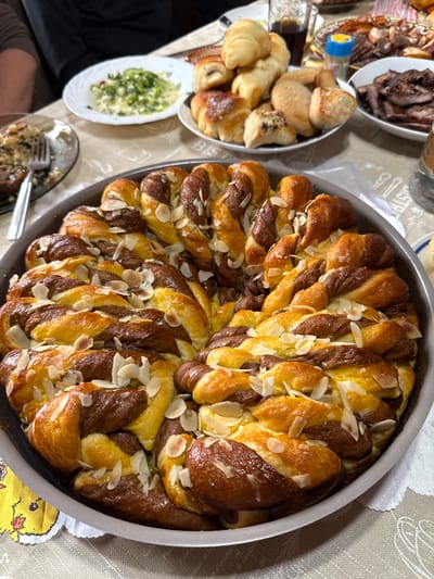 The image captures a dining table setting with various dishes, possibly in a home in Vinarovo, Bulgaria. A round braided bread, topped with sliced almonds, occupies the center of the frame, sitting in a gray baking pan. Other dishes, including pastries, sliced meat, and a salad with a creamy dressing, are arranged around the bread. The tablecloth is light-colored with a subtle pattern. A fork rests on a plate in the lower-left corner of the frame, indicating ongoing or recent dining activity.
