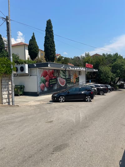 The image depicts a sunny day scene in Crikvenica, Croatia, featuring a Konzum supermarket. In the foreground, a paved parking lot with asphalt is visible, with three dark-colored cars parked in a row. The cars are a black Volkswagen Golf, a dark grey SUV, and a black sedan. To the left of the parking lot, a low stone wall separates the lot from a concrete sidewalk. A green dumpster is situated on the sidewalk next to the supermarket building.
The supermarket itself has a modern, somewhat weathered appearance, with a dark-colored awning above the entrance. The front of the store is adorned with large photographic murals of fresh produce, including raspberries, watermelon, and apricots. Text in Croatian is visible on the murals, such as "OD POLJA DO STOLA" (From field to table) and "UVIJEK KUPUITE DOMACE!" (Always buy local!). Above the awning, the red "KONZUM" logo is prominently displayed. Air conditioning units are mounted on the exterior wall of the building.
In the background, several residential buildings with red tile roofs and a dense line of trees, including tall cypress trees, are visible. The sky is clear and bright blue with a few scattered white clouds. A utility pole with electrical wires is also present on the left side of the image. The overall impression is a typical Mediterranean commercial area on a bright, warm day.