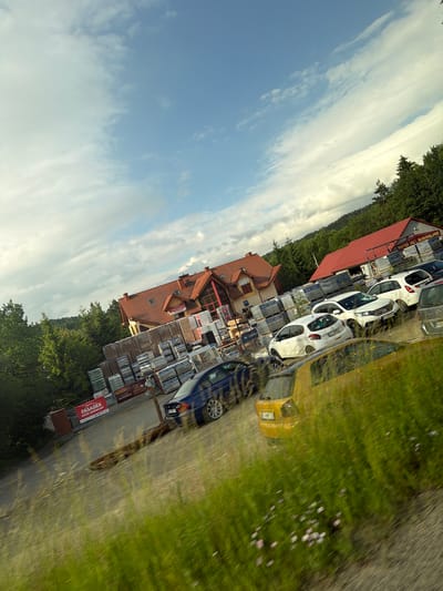 The image depicts an outdoor view of a building materials store with a parking lot filled with cars. The main building has a red tiled roof and is surrounded by stacks of building materials, including what appear to be pallets of bricks or stone. Several cars are parked in the lot, including a blue sedan, a yellow hatchback, and several white cars. The scene is set against a backdrop of trees and a partially cloudy sky. A sign is visible on the left side of the image that reads "FASADEK SKŁAD BUDOWLANY". Based on this sign, the location is likely a building materials store in Limanowa, Poland. The image is taken from a slightly elevated angle and appears to be from a moving vehicle, as indicated by the motion blur in the foreground vegetation and some of the cars.