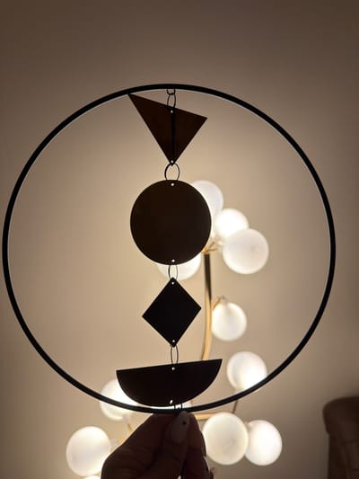 An indoor shot shows a decorative mobile hanging in front of a ceiling light fixture. The mobile is circular with a black frame and contains several geometric shapes suspended from it by chains. These shapes include a triangle, a circle, a diamond, and a semi-circle, all made of dark material and silhouetted against the light. The background reveals a cluster of glowing white orbs from a chandelier-like light fixture. A hand with manicured nails is visible at the bottom, holding the mobile, suggesting someone is interacting with it. The lighting creates a warm, diffused ambiance. The scene is set in Mojanovići, Montenegro.