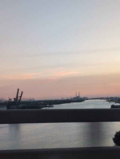 The image shows a wide view of a harbor area in Oudalle, France, during what appears to be either sunrise or sunset. The sky is a gradient of soft colors, transitioning from pale blue at the top to pale pink and orange near the horizon, with faint wisps of clouds.
In the foreground, a dark, blurred horizontal structure, likely a railing or barrier, divides the image. Below this, a body of water, possibly a canal or river, reflects the colors of the sky. The water's surface is calm, with subtle ripples.
The midground reveals a busy industrial port. Prominent features include large cranes with tall, skeletal structures, characteristic of port loading equipment. Several ships or boats are visible on the water. In the distance, industrial buildings and structures, including what appear to be power plant smokestacks, dot the landscape. Lights are visible along the waterfront, suggesting ongoing activity despite the low light.
The overall atmosphere is one of industrial activity set against a tranquil, colorful sky. The precise location is identified as the city of Oudalle, France, known for its port facilities. There are no discernible people or direct human interactions depicted in the image. The focus is on the vast scale of the industrial infrastructure and its natural surroundings.