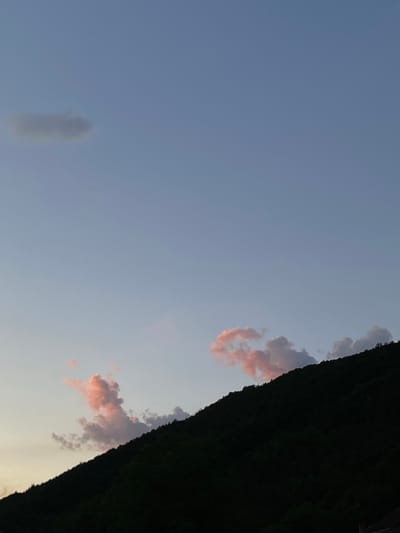 The image shows a dusky sky over a dark, tree-covered hillside. Two wisps of cloud in the upper left are tinged grey, while two others near the center of the frame are illuminated with a soft pink hue from the setting sun. The hillside occupies the lower right portion of the frame, its dark silhouette obscuring any details of the terrain or any potential structures. The sky is a pale blue, transitioning to a lighter shade near the horizon, indicating the twilight hour. There are no people or discernible human activity in the frame. The scene evokes a sense of quietude and natural beauty typical of a rural landscape at dusk. This scene could be from Prijepolje Municipality, Serbia, known for its mountainous terrain and natural landscapes.