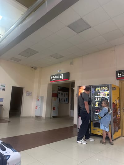 In Lyambir', Russia, a young girl in a light blue denim dress and jacket is reaching into an M&M's vending machine. She appears to be contemplating her purchase. Beside her, a man in a dark grey t-shirt and black pants, wearing a black baseball cap, stands with his arm around her, also looking at the machine. He has his eyes closed, and his head is tilted slightly, suggesting a moment of quiet intimacy or shared enjoyment.
The setting appears to be an indoor public space, possibly a shopping mall, airport, or train station, indicated by the tiled floor, suspended ceiling tiles, and illuminated exit signs in both Russian and English. One sign clearly reads "Выход Exit". Other signs and fixtures suggest a commercial or service area, including information boards and what looks like other vending machines or service points. The lighting is bright and artificial, suggesting it could be daytime or evening. There are no specific indications of the weather. The overall mood seems relaxed and casual, focused on the interaction between the two individuals and the vending machine.