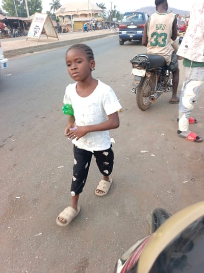 A young girl stands in a street, wearing a white shirt, cropped black pants with white spots, and light-colored sandals. She holds a green object in her left hand and a purple object in her right. Behind her, a man on a motorcycle wears a "Bird 33" jersey. A blue van is parked nearby. A sign is visible on the left, and other people and buildings line the street in the background.