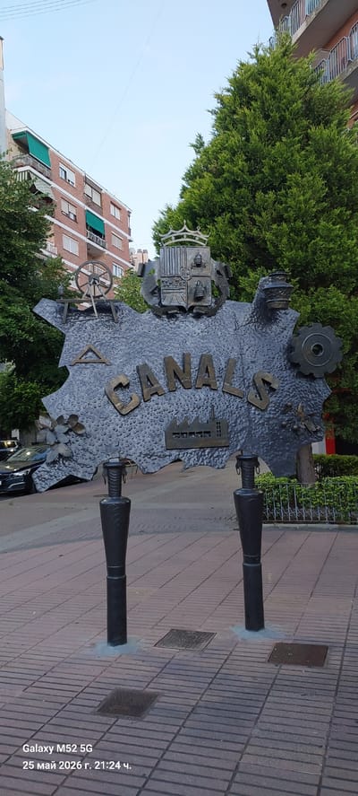 A metal sculpture representing the town of Canals in Spain, sits on a brick-paved sidewalk. The word "CANALS" is prominently displayed on the sculpture, accompanied by various metal cutouts including a gear, coat of arms, factory, a wheel, and flowers. The sculpture is supported by two cylindrical metal posts. Behind the sculpture are a green hedge, a street, a multi-story brick building with awnings, and a sky with some visible clouds. The scene is located in the city of l'Alcúdia de Crespins, Spain. The date and time "25 May 2026 г. 21:24 ч." are printed in the lower right corner.