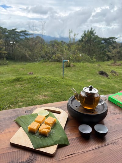 The image shows a tea set and some snacks placed on a wooden table outdoors, with a view of a lush green landscape under a cloudy sky. The scene appears to be set in Ban Khun Wang, Thailand, given the surrounding vegetation which is typical of the region. In the foreground, a clear glass teapot filled with amber-colored tea sits on a black circular stand. Next to it are two small, dark-rimmed ceramic cups. To the left, on a wooden cutting board topped with a banana leaf, are four square pieces of light orange confectionery, possibly fudge or a similar sweet, dusted with white powder. A slice of lemon and a small cinnamon stick rest on the banana leaf. In the background, the landscape features rolling hills covered in trees, with a visible pathway or clearing. The sky is overcast with large, fluffy clouds, suggesting a potentially cool or humid day. The overall atmosphere is one of a tranquil, outdoor tea-time experience.