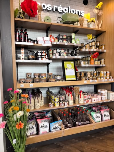 This is a medium-shot, eye-level interior photograph of a shelf display in a retail store. The shelves are stocked with various food products, primarily in jars and bottles, and packaged goods.

The top shelf displays decorative items, including a red poppy, a golden Eiffel Tower replica, and a small green cart with the text "HELLO SPRING" on it. Several jars of preserves and condiments are also present.

The second shelf from the top holds more jars of preserves and condiments, as well as some packaged food items.

The third shelf contains several rows of small jars, likely containing jams, honey, or spreads.

The fourth shelf is filled with jars and bottles, including what appear to be mustard jars and other condiments.

The bottom shelf has a prominent display of bagged charcoal briquettes, some with checkered patterns, and several bottles of liquid.

In the foreground, on the left side of the image, a bouquet of artificial flowers in shades of pink, orange, and white is visible.

The store appears to be well-lit, with a focus on the merchandise. The backdrop is a white tiled wall. The overall impression is a well-organized retail environment showcasing regional food products. The store is located in Briis-sous-Forges, France.