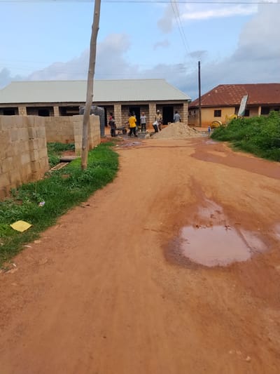 The image depicts a construction site in Mangu Town, Nigeria, with several individuals engaged in work. In the foreground, a dirt road occupies the majority of the frame, with a large puddle of muddy water reflecting the sky. To the left of the road, a partially constructed building made of concrete blocks is visible, with a water tank nearby. Several people are working on this structure, some carrying materials and others appearing to be mixing cement. Further into the scene, another building with a corrugated metal roof is under construction. In the background, a second building with a reddish-brown metal roof stands, and a yellow cement mixer is present. Lush green vegetation borders the right side of the frame. Power lines are strung across the sky, supported by a wooden pole and a utility pole. The overall impression is one of ongoing construction work in a semi-urban environment.