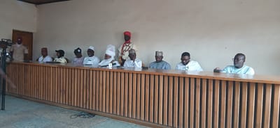A group of men and one woman are seated at a long, wooden table in what appears to be a meeting room. The room has a neutral-colored wall and a wooden ceiling. The table is fronted by a wooden lattice structure.

There are at least nine individuals visible. From left to right, one person in a light brown uniform stands near a doorway. Seated at the table are several men wearing traditional attire, including caps and robes. One individual in the center is wearing a prominent white turban and elaborate robes, possibly indicating a significant role. Another person to the right of the turbaned individual is wearing glasses and a light blue shirt. The remaining individuals are also dressed in traditional clothing, mostly in white. Some have microphones in front of them.

To the left of the table, a tripod with a camera or recording device is visible. The floor appears to be tiled. The overall scene suggests a formal gathering or a presentation. The presence of the uniform suggests an official capacity. Given the mention of Sabon Gari North, Nigeria, the setting is likely a community hall or government building within that region.
