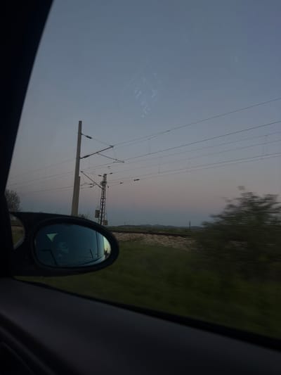 The image shows a view from inside a vehicle, likely a car, with a side mirror visible. The scene outside the car depicts a landscape with power lines stretching across the sky. The sky appears to be a gradient of light blue to a pale orange-pink, possibly indicating sunrise or sunset. There is some vegetation visible to the right of the frame, along with what appears to be a railway track. The location is likely rural, suggested by the presence of power lines and greenery. While there is no visual confirmation of the Dalgopol, Bulgaria location, it can be inferred from the presence of railway lines and power lines, infrastructure commonly found in populated areas. No people are visible in the image. The reflection in the side mirror is unclear, but shows the presence of a vehicle. The action is movement, implied by the blurred scenery.