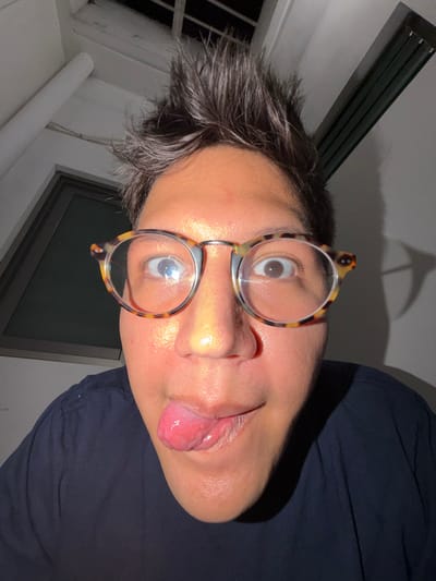An individual, wearing round eyeglasses, faces the camera with their head tilted. Their tongue is extended. Dark hair appears disheveled. The person is wearing a dark-colored shirt. Light shines from above, casting a shadow in the background. A window is visible in the lower portion. A light fixture with horizontal slats is partially visible in the upper left.
