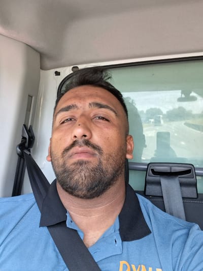 A man, appearing to be of Middle Eastern descent, is pictured from the chest up, seated in the passenger seat of a vehicle. He has dark hair, a prominent nose, a full beard, and is wearing a light blue collared shirt with a black collar and a black seatbelt across his chest. The background shows the interior of a vehicle with a seatbelt buckle visible, and through the rear window, a highway scene with trees and other vehicles can be observed. The image was taken in the city of Londerzeel, Belgium.