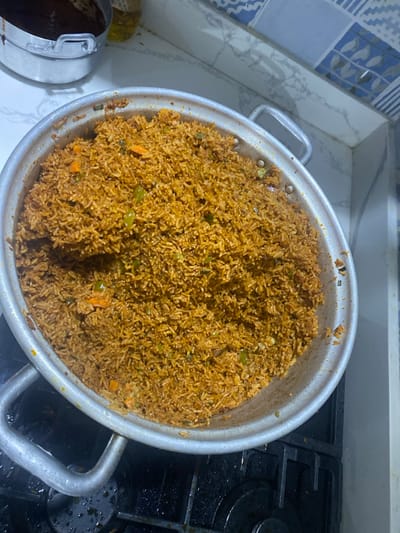 A large metal pot brimming with cooked Jollof rice is the central subject of this overhead shot, likely taken in a kitchen in Adenta Municipality, Ghana. The rice displays a characteristic reddish-brown hue, interspersed with visible pieces of chopped carrots and green bell peppers. The pot itself is silver-colored, with two sturdy handles, suggesting it's a sizable vessel for cooking large quantities.
The pot is situated on a kitchen surface, with the left side resting on a white, marble-patterned countertop. A blue and white patterned tiled backsplash is visible behind this section of the counter. The right side of the pot extends over a black gas stovetop, which has visible grates and some minor cooking residue, indicating it has been recently used. A portion of another metallic pot or container is faintly visible on the far left of the image. The scene conveys the preparation of a hearty meal, typical of home cooking. There is no visible text in the image.