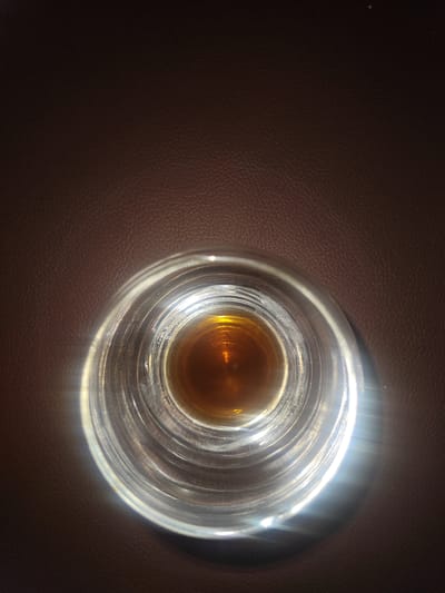 An overhead shot shows a glass containing amber liquid on a brown textured surface. The glass is partially illuminated from the right side, creating a bright glare. The liquid is a warm, dark amber color. The brown surface appears to be leather with a visible grain pattern. The scene is devoid of any people or city landmarks. This image is not of Modena, Italy.