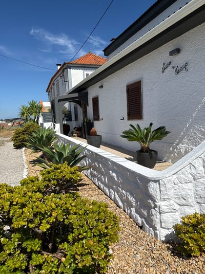 The image depicts the exterior of a modern white building with dark grey accents and an orange tiled roof, located in Almada, Portugal. The building features dark brown shutters on windows and black decorative script on the exterior wall that reads "Nuno e Tiago". In the foreground, a low white stone wall borders a garden with various plants. These include several large agave plants, a bushy green succulent, and a palm plant in a dark pot. The ground is covered with small, light-colored gravel. Behind the main building, another structure with an orange roof and white walls is partially visible. The sky is clear blue with a few wispy white clouds. The scene suggests a sunny day in a residential or commercial area with a focus on landscaping.
