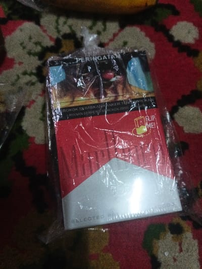 A retail-packaged Marlboro cigarette carton, still encased in its clear plastic wrapper, rests centrally on a patterned textile surface. The textile features a predominantly red background with abstract white, green, and dark brown motifs. The cigarette carton displays the characteristic red and white Marlboro design. On the upper portion of the packaging, a graphic health warning is visible, featuring an image of diseased human lungs and text in Indonesian: "PERINGATAN" (Warning) and "ROKOK SEBABKAN KANKER TENGGOROKAN" (Smoking Causes Throat Cancer). Below this warning, a "FLIP ME!" prompt with an associated icon is present on the red section of the carton. The phrase "SELECTED PREMIUM TOBACCOS" appears near the bottom edge. A blurry, yellowish-brown object is partially visible in the upper background. No individuals are present in the scene, which is situated indoors in Kon Tum, Vietnam.