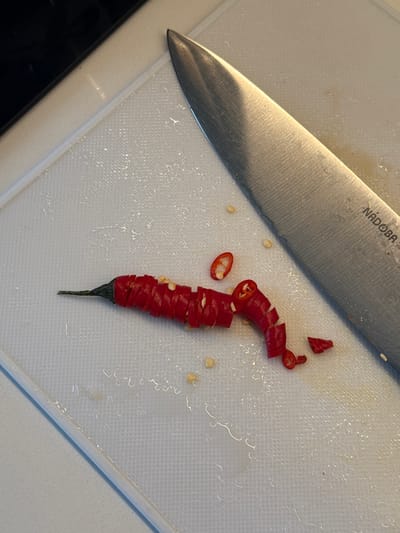 An overhead close-up shot depicts a culinary preparation scene in what appears to be a kitchen. The primary subjects are a large chef's knife and a sliced red chili pepper, both resting on a white plastic cutting board.

The metallic chef's knife is positioned diagonally across the top left quadrant of the frame, with its blade angled towards the lower center. The brand name "NADOBA" is clearly visible, etched into the upper part of the blade. Below "NADOBA," partially obscured text that seems to read "chef" or similar is also present. The blade shows signs of use with slight scratches and reflections from ambient light.

To the right of the knife, a red chili pepper has been uniformly sliced into numerous thin, bright red rings. These slices are arranged in a stack, largely preserving the original form of the pepper, with its dark green stem still intact at the bottom. Several pale yellow chili seeds are scattered both among the pepper slices and across the textured surface of the cutting board. A few smaller, irregularly shaped chili pieces are also visible.

The white cutting board features a distinctive textured, grid-like pattern across its surface. Small droplets of liquid or moisture are present on the board, particularly around the sliced pepper, suggesting recent activity. A dark edge, likely a countertop or kitchen surface, is visible along the bottom and left side of the cutting board. The scene captures a functional moment of food preparation in Shirkovo, Russia.